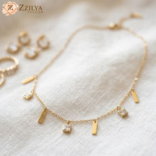 Gold charms necklace with square and bar pendants on a white fabric background, branded 'ZZILYA'.