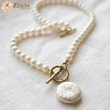 Close-up of a ZZILYA Fashion pearl necklace featuring a strand of round white pearls, an antique gold-finish toggle clasp, and a flat white coin pearl pendant resting on a white textured fabric background.
