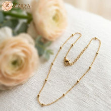 Delicate gold-toned satellite chain necklace with small polished beads by Zzilya Fashion, displayed on a white fabric surface next to soft peach ranunculus flowers.