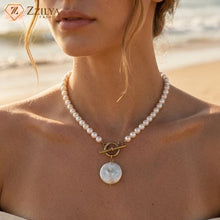 Close-up of a ZZILYA Fashion pearl necklace featuring a strand of round white pearls, an antique gold-finish toggle clasp, and a flat white coin pearl pendant resting on a white textured fabric background.
