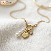 Gold necklace with heart and pearl charm on a white fabric background, branded 'Zzilya'.
