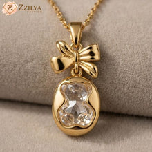 Gold pendant with bow design and clear gemstone on a beige background, branded 'Zzilya'.