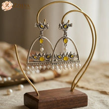 Ornate earrings with yellow stones and pearl-like beads on a wooden stand, brand 'Zzilya' visible.