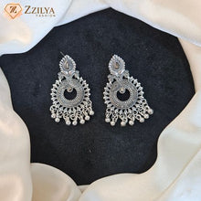 A pair of ornate silver-toned Indian-style dangle earrings with intricate filigree work and small bead drops, displayed on a black textured insert surrounded by draped white fabric. The brand logo 'Zzilya Fashion' is visible in the top left corner.