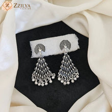 A pair of ornate silver-toned Indian peacock earrings with detailed filigree and dangling bead chains, displayed on a black textured surface with a white fabric backing and draped cream satin fabric by zzilya fashion