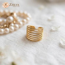Gold ring on a textured white surface with blurred pearls in the background, featuring the brand 'Zzilya'.