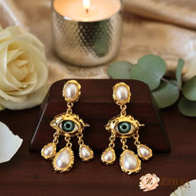 ZZILYA Fashion gold chandelier earrings featuring a central green evil eye motif and multiple hanging white teardrop pearls on a wooden display.