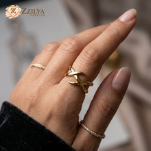 Close-up of a person's hand showcasing three stacking rings: a bold golden X-shaped crossover ring on the middle finger, paired with two delicate cubic zirconia eternity bands on the index and pinky fingers, set against a blurred textile background with the ZZILYA Fashion logo.
