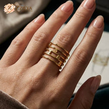 Close-up of a person's hand wearing a unique golden multi-layer stacker ring on the ring finger. The ring is composed of five thin, connected bands, and features four small, circular gold discs, set on various levels. The background shows blurred textiles and a sketch, with the ZZILYA Fashion logo in the corner.