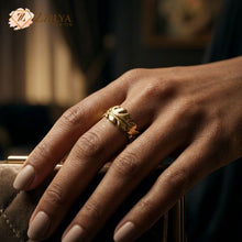 Close-up of a hand with nude manicured nails wearing a distinct gold feather ring, with a texture and design resembling a leaf or feather, resting gently on a brown textured object by Zzilya fashion Jewellery