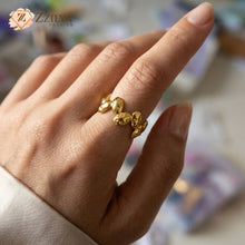 Close-up of a hand with nude nails wearing a unique, segmented gold ring composed of several interconnected, irregular polished gold shapes, resembling abstract nuggets or links. The hand is in a relaxed gesture, set against a softly blurred desk background with color swatches and art supplies, and features a ZZILYA FASHION logo.