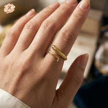 A close-up of a hand with a natural manicure wearing a textured gold snake ring that wraps around the ring finger. The ring features intricate scale detailing and a serpent head design. The background is softly blurred with a hint of a jewelry box and the ZZILYA Fashion logo.