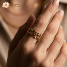 A high-angle shot of a polished golden chain-link style ring resting on a textured off-white fabric. The ring features an open, adjustable back. In the blurred background, matching gold and pearl jewelry are visible, with the ZZILYA Fashion logo in the top-left corner.