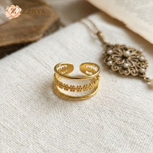 An adjustable gold-toned triple-layer ring resting on a white textured fabric. The ring features two smooth outer bands and a center band with a detailed, beaded texture. A vintage pearl necklace is visible in the soft-focus background by zzilya Fashion