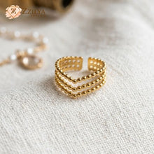 A close-up shot of a ZZILYA 18K gold-plated open ring featuring three stacked rows of gold beads in a chevron V-shape, displayed on a white textured fabric background.