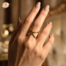 woman’s hand wearing a textured gold X-shaped statement ring on her middle finger, showcasing a hammered metal finish against a warm, blurred background By Zzilya Fashion Jewelery