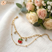 ZZILYA Fashion double-layered gold charm bracelet featuring a red enamel cherry pendant and bezel-set crystals, displayed on a white linen fabric next to pale pink roses.