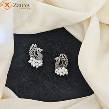 A pair of silver-toned peacock stud earrings with white pearl clusters, displayed on a black textured card surrounded by draped cream fabric. The brand logo 'Zzilya Fashion' is visible in the top left corner.