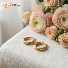 Gold hoop earrings on a white surface with pink flowers in the background, featuring the brand 'Zzilya'.