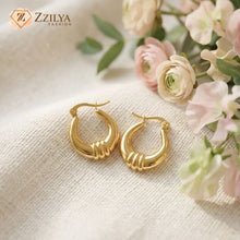 Gold hoop earrings on a textured surface with flowers in the background, featuring the brand 'ZZILYA'.