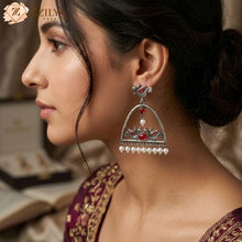 Close-up product shot of Zzilya Fashion's trendy silver oxidized earrings featuring an arch shape decorated with traditional peacock and floral motifs, inlaid with deep teal stones, and finished with a neat row of white hanging pearls resting on a textured linen background.