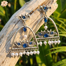 Ornate silver earrings with blue stones and pearls on a wooden branch with green leaves in the background by zzilya fashion