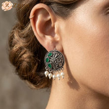 Close-up of an ear wearing a decorative earring with green gemstone and pearl drops, brand 'Zzilya' visible.