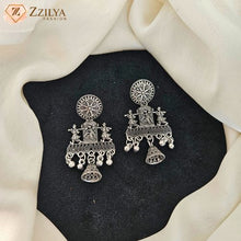 A pair of ornate silver-toned Indian-style dangle earrings with intricate filigree and bell-shaped drops, displayed on a black textured surface surrounded by draped cream fabric. The Zzilya Fashion logo is visible in the top-left corner.