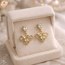 Gold earrings with clear stones on a beige jewelry box, branded 'ZZILYA'.