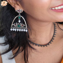 Close-up of a woman wearing silver earrings with gemstones and pearls, branded 'Zyilya'.