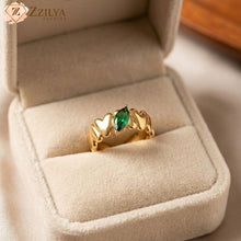 A golden anti-tarnish adjustable ring by Zzilya Fashion featuring a band of gold hearts and a central marquise-cut green emerald zircon, displayed in a plush beige jewelry box.