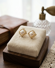 Pair of square-shaped gold stud earrings featuring a raised mother-of-pearl center and a geometric pavé crystal border by Zzilya Fashion.