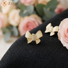 Gold bow-shaped earrings on a black surface with pink flowers in the background