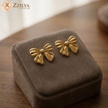 Gold bow-shaped earrings on a brown velvet cushion with 'Zzilya Fashion' branding.