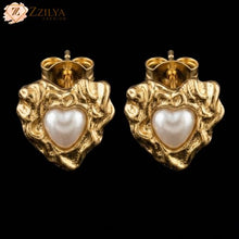 Gold heart-shaped earrings with a pearl center on a black background, branded 'ZZILYA'.