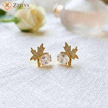 Gold maple leaf-shaped earrings with clear stones on a textured white surface, branded 'Zzilya'.