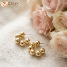 Gold hoop earrings on a textured surface with pink roses in the background, featuring the brand 'Zzilya'.