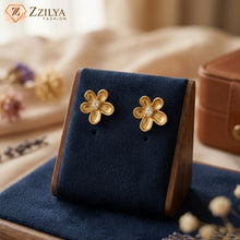 Gold flower-shaped earrings on a blue stand with 'ZZILYA' branding.