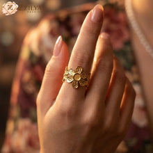 Gold flower ring on a hand with a blurred floral background, featuring the brand 'ZZILYA'.