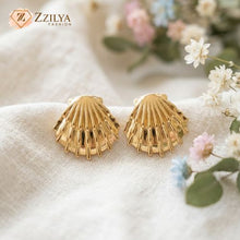Gold shell-shaped earrings on a textured surface with flowers in the background, featuring the brand 'ZZILYA'.