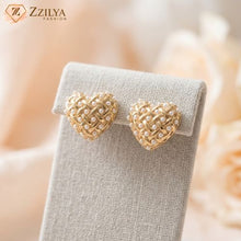 Heart-shaped gold earrings on a gray stand with a soft blurred background