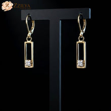 Gold earrings with rectangular design and clear stones on a black stand, branded 'Zzilya'.
