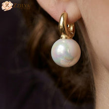 Pearl earring with gold setting worn by a person, brand 'ZZILYA' visible.