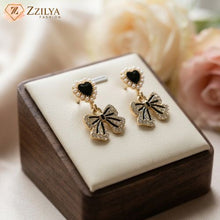 Pair of gold and black bow-shaped earrings on a wooden box with a soft floral background. by Zzilya fashion