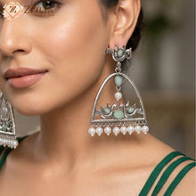 Close-up of a woman wearing ornate silver earrings with green stones and pearls, branded 'Zzilya'.