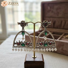 Close-up product shot of Zzilya Fashion's trendy silver oxidized earrings featuring an arch shape decorated with traditional peacock and floral motifs, inlaid with deep teal stones, and finished with a neat row of white hanging pearls resting on a textured linen background.