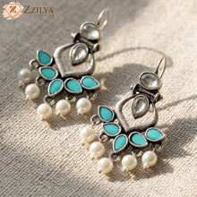 Pair of silver earrings with turquoise stones and pearls on a textured surface, branded 'Zzilya'.