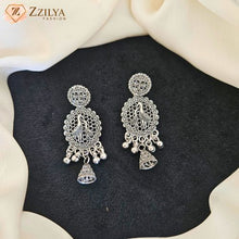 A pair of ornate silver-toned Indian jhumka earrings with intricate filigree work, peacock motifs, and dangling bell charms, displayed on a black textured surface with draped cream fabric.