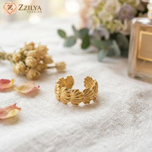 Golden seashell ring on a textured surface with flowers and a perfume bottle in the background, featuring the brand 'Zzilya'.