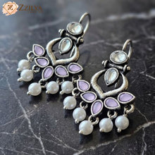 Silver earrings with pearls and purple stones on a dark stone surface, branded 'Zzilya'.
